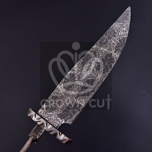 May include: A large, ornate blade with a floral pattern etched into the steel. The blade has a wide, pointed tip and a cross-shaped handle. The metal has a dark, matte finish, and the floral design is in a lighter tone, creating a striking contrast.