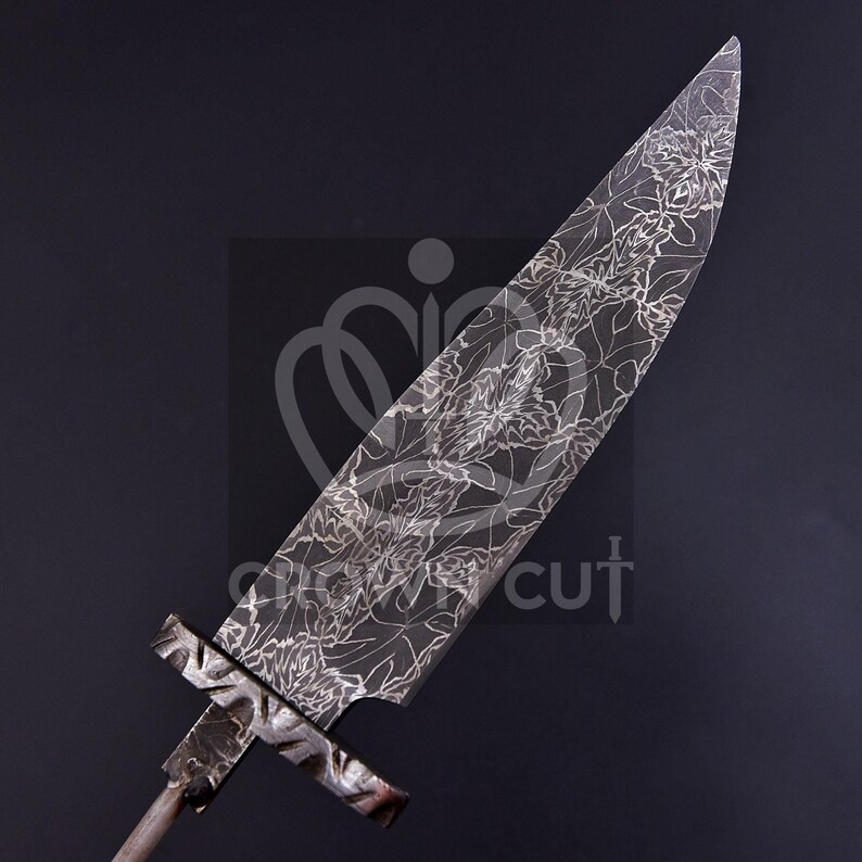 May include: A large, ornate blade with a floral pattern etched into the steel. The blade has a wide, pointed tip and a cross-shaped handle. The metal has a dark, matte finish, and the floral design is in a lighter tone, creating a striking contrast.