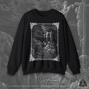 May include: Black crewneck sweatshirt featuring a detailed black and white illustration of a religious scene. The artwork, framed with an ornate border, depicts figures around three crosses under a dramatic sky. The text "THE CRUCIFIXION" is at the bottom of the image.