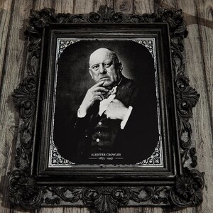 May include: A black and white portrait of Aleister Crowley, 1875-1947, encased in an ornate, dark, carved rectangular frame. The image features a man in a suit with his hand near his chin, set against a dark background with white floral corner accents.
