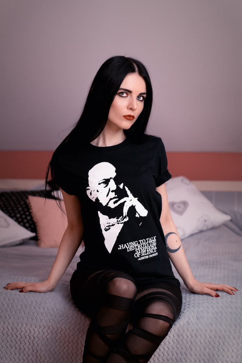 Puede incluir: Una mujer con el pelo largo y negro lleva una camiseta negra con una imagen blanca de un hombre y el texto "Having to talk destroys the symphony of silence - Aleister Crowley". Est&aacute; sentada en una cama con un tatuaje de luna creciente en el brazo.