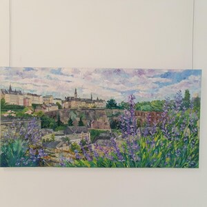 Puede incluir: Una pintura de paisaje que representa una ciudad europea con edificios y un cielo nublado. Flores moradas en primer plano. La obra de arte se exhibe en una pared blanca, suspendida por un cable.