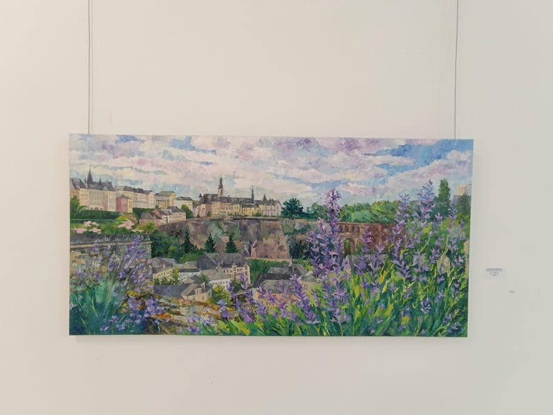 Puede incluir: Una pintura de paisaje que representa una ciudad europea con edificios y un cielo nublado. Flores moradas en primer plano. La obra de arte se exhibe en una pared blanca, suspendida por un cable.