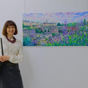 Puede incluir: Una pintura al &oacute;leo de un paisaje urbano con una paleta de colores vibrantes. La obra de arte representa edificios, &aacute;rboles y un cielo nublado, con flores moradas en primer plano. La pintura se exhibe en una pared blanca.