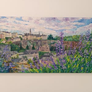 Puede incluir: Pintura al &oacute;leo de un paisaje urbano europeo con edificios, un puente y flores moradas en primer plano. El cielo es una mezcla de azul y blanco, y la pintura est&aacute; sobre un lienzo rectangular.