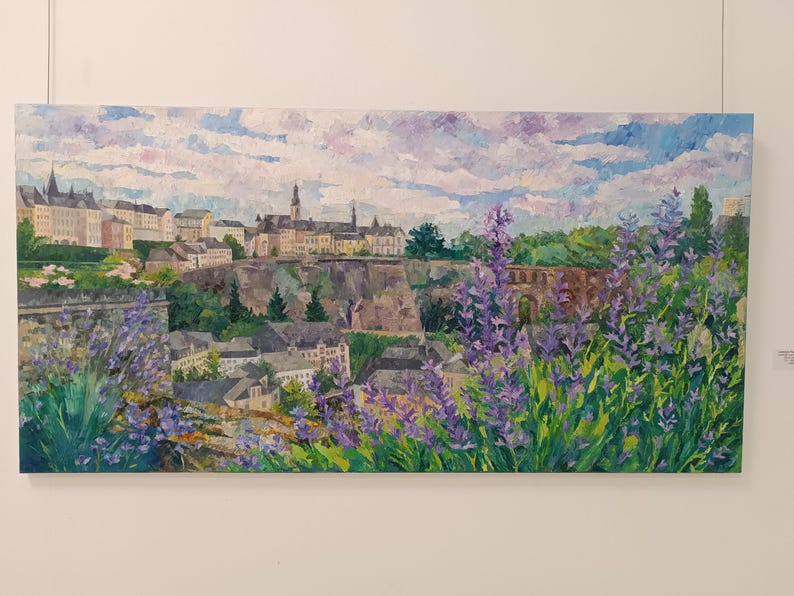 Puede incluir: Pintura al &oacute;leo de un paisaje urbano europeo con edificios, un puente y flores moradas en primer plano. El cielo es una mezcla de azul y blanco, y la pintura est&aacute; sobre un lienzo rectangular.