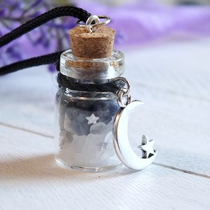 Sea Salt And Blue Sodalite Chips in a small Spell Bottle With Crescent Moon Charm Necklace on white wooden surface with purple flowers and green leaves in the background at top.