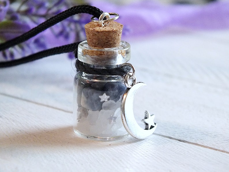 Sea Salt And Blue Sodalite Chips in a small Spell Bottle With Crescent Moon Charm Necklace on white wooden surface with purple flowers and green leaves in the background at top.