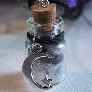 Sea Salt And Blue Sodalite Chips in a small Spell Bottle With Crescent Moon Charm Necklace on white wooden surface with purple flowers and green leaves in the background at top.