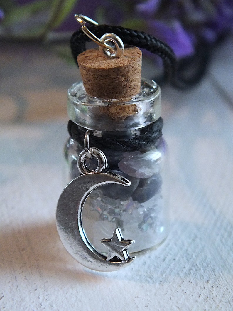 Sea Salt And Blue Sodalite Chips in a small Spell Bottle With Crescent Moon Charm Necklace on white wooden surface with purple flowers and green leaves in the background at top.