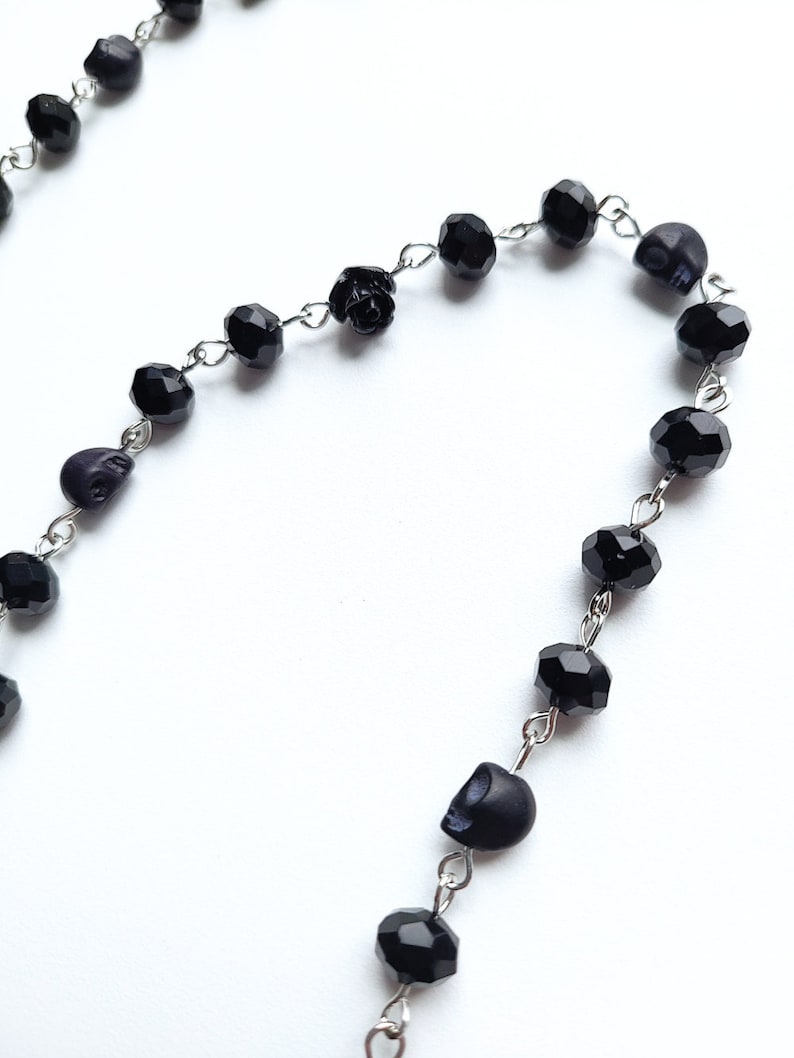 May include: A black beaded necklace with skull and rose charms. The necklace is made of silver chain and black beads.