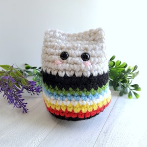 May include: A crocheted cat toy with a white, black, green, blue, yellow, and red striped body. The cat has black eyes and pink cheeks.