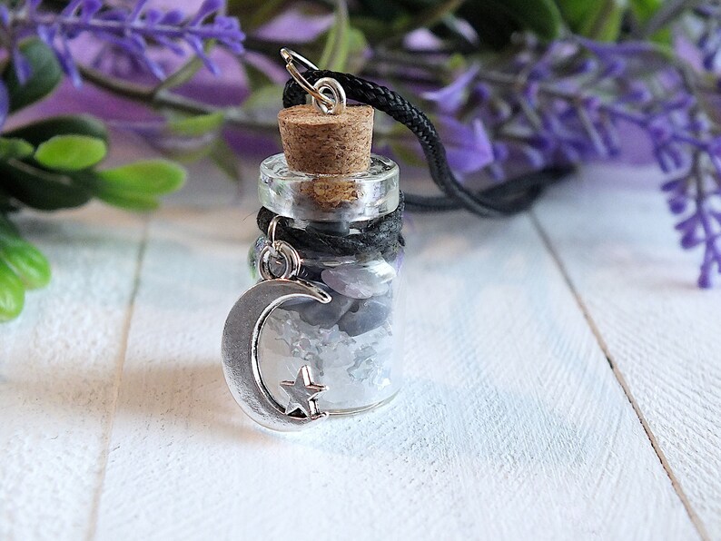 Sea Salt And Blue Sodalite Chips in a small Spell Bottle With Crescent Moon Charm Necklace on white wooden surface with purple flowers and green leaves in the background at top.