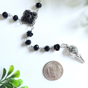 May include: Black beaded necklace with a silver skull charm and a silver filigree pendant. The necklace also features black rose accents.