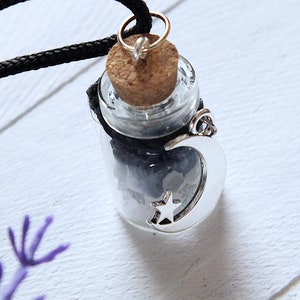 Sea Salt And Blue Sodalite Chips in a small Spell Bottle With Crescent Moon Charm Necklace on white wooden surface with purple flowers at the bottom.