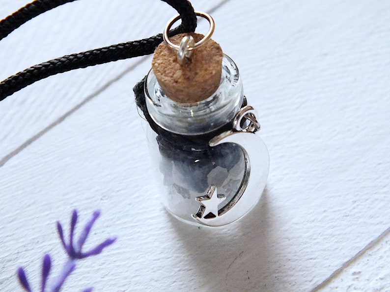 Sea Salt And Blue Sodalite Chips in a small Spell Bottle With Crescent Moon Charm Necklace on white wooden surface with purple flowers at the bottom.