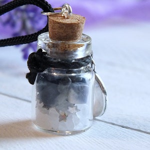 Sea Salt And Blue Sodalite Chips in a small Spell Bottle With Crescent Moon Charm Necklace on white wooden surface with purple flowers and green leaves in the background at top.