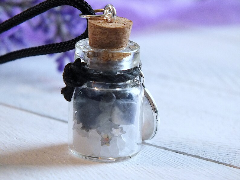 Sea Salt And Blue Sodalite Chips in a small Spell Bottle With Crescent Moon Charm Necklace on white wooden surface with purple flowers and green leaves in the background at top.