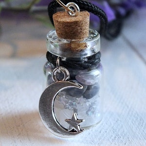 Sea Salt And Blue Sodalite Chips in a small Spell Bottle With Crescent Moon Charm Necklace on white wooden surface with purple flowers and green leaves in the background at top.