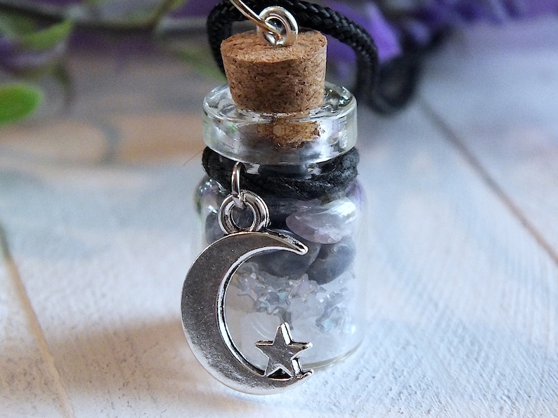 Sea Salt And Blue Sodalite Chips in a small Spell Bottle With Crescent Moon Charm Necklace on white wooden surface with purple flowers and green leaves in the background at top.