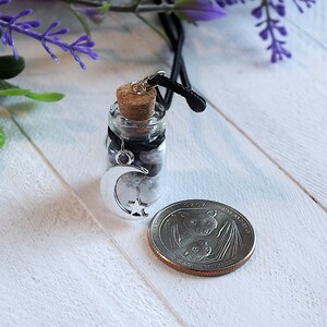 Sea Salt And Blue Sodalite Chips in a small Spell Bottle With Crescent Moon Charm Necklace on white wooden surface with purple flowers and green leaves in the background at top and a quarter next to it for size.