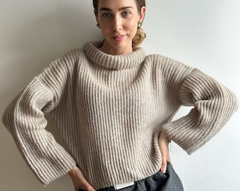Könnte beinhalten: Ein beiger Rollkragenpullover mit gerippter Struktur und langen Ärmeln. Der Pullover ist kurz geschnitten und hat eine lockere Passform. Das Model trägt den Pullover zu einer grauen Hose.