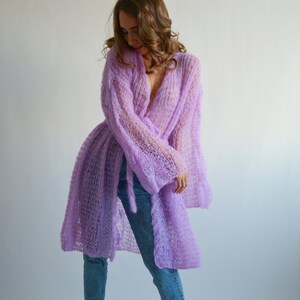 May include: A long, light purple knitted cardigan with a loose fit and a tie belt. The cardigan is made of a delicate, open-weave fabric.