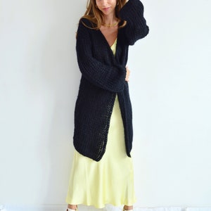 Op de afbeelding: Een vrouw draagt een lange zwarte gebreide cardigan over een gele satijnen slipdress. Ze draagt gouden sieraden en heeft haar haar naar achteren getrokken.