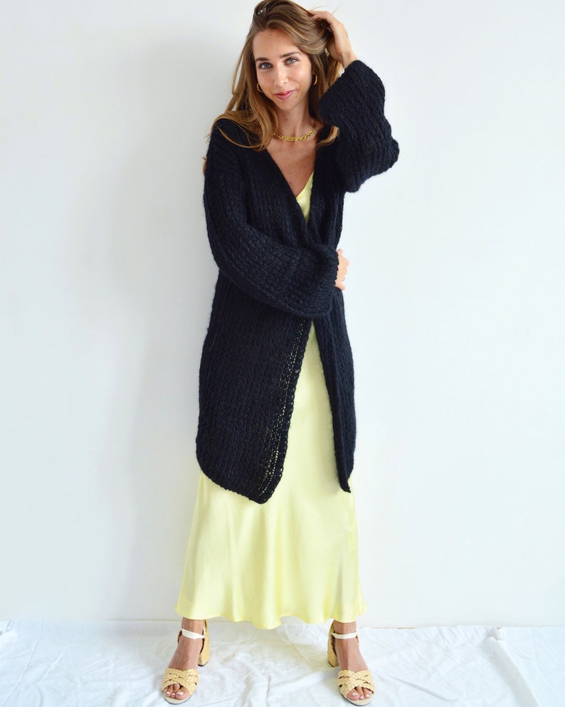 Op de afbeelding: Een vrouw draagt een lange zwarte gebreide cardigan over een gele satijnen slipdress. Ze draagt gouden sieraden en heeft haar haar naar achteren getrokken.
