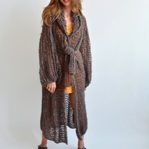May include: A long, brown, open-front cardigan with a tie belt. The cardigan is made of a loose knit fabric with a speckled pattern. The model is wearing the cardigan over a yellow tank top and black sandals.