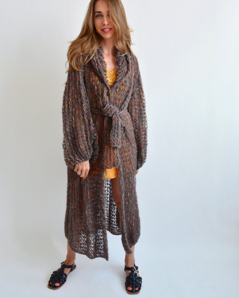 May include: A long, brown, open-front cardigan with a tie belt. The cardigan is made of a loose knit fabric with a speckled pattern. The model is wearing the cardigan over a yellow tank top and black sandals.
