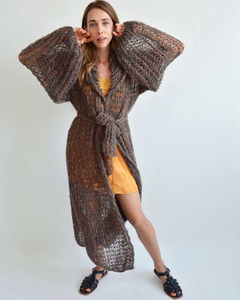 May include: A long, brown, knit cardigan with a tie belt and large sleeves. The cardigan has a loose, flowing fit and is made of a soft, textured yarn.