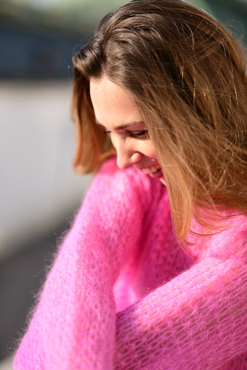 以下が含まれることがあります： ピンクのふわふわのセーターを着た女性が下を見て微笑んでいます