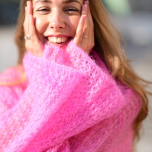 以下が含まれることがあります： ゆったりとしたピンクのふわふわのセーターを着た女性。彼女は笑顔で、両手で頬を触っています。