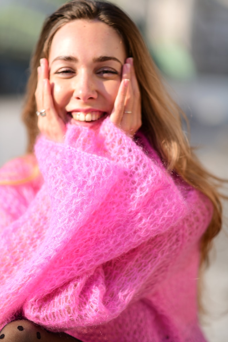 以下が含まれることがあります： ゆったりとしたピンクのふわふわのセーターを着た女性。彼女は笑顔で、両手で頬を触っています。