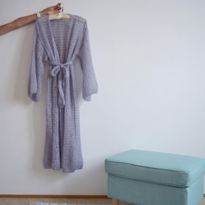 May include: A light purple knitted cardigan with long sleeves and a tie belt. The cardigan is hanging on a wooden hanger against a white wall. A light blue footstool is in the foreground.