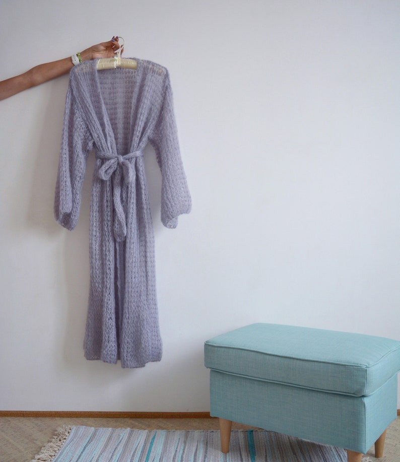 May include: A light purple knitted cardigan with long sleeves and a tie belt. The cardigan is hanging on a wooden hanger against a white wall. A light blue footstool is in the foreground.