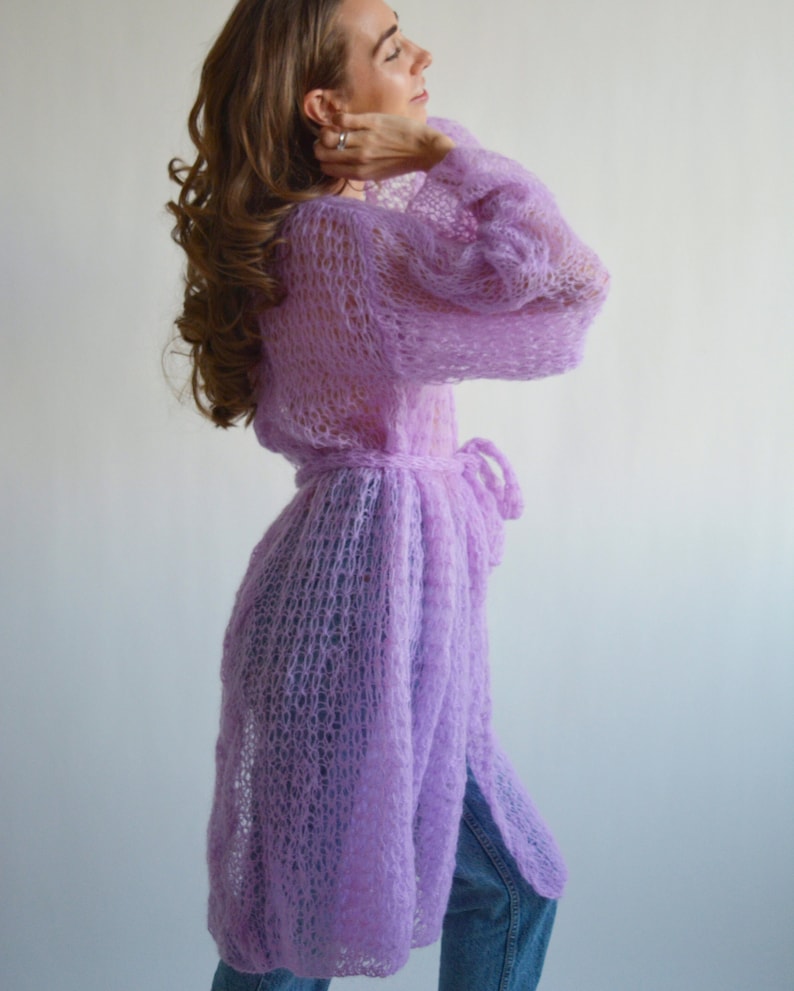May include: A long, light purple knitted cardigan with a tie belt. The cardigan is made of a loose, open knit fabric.