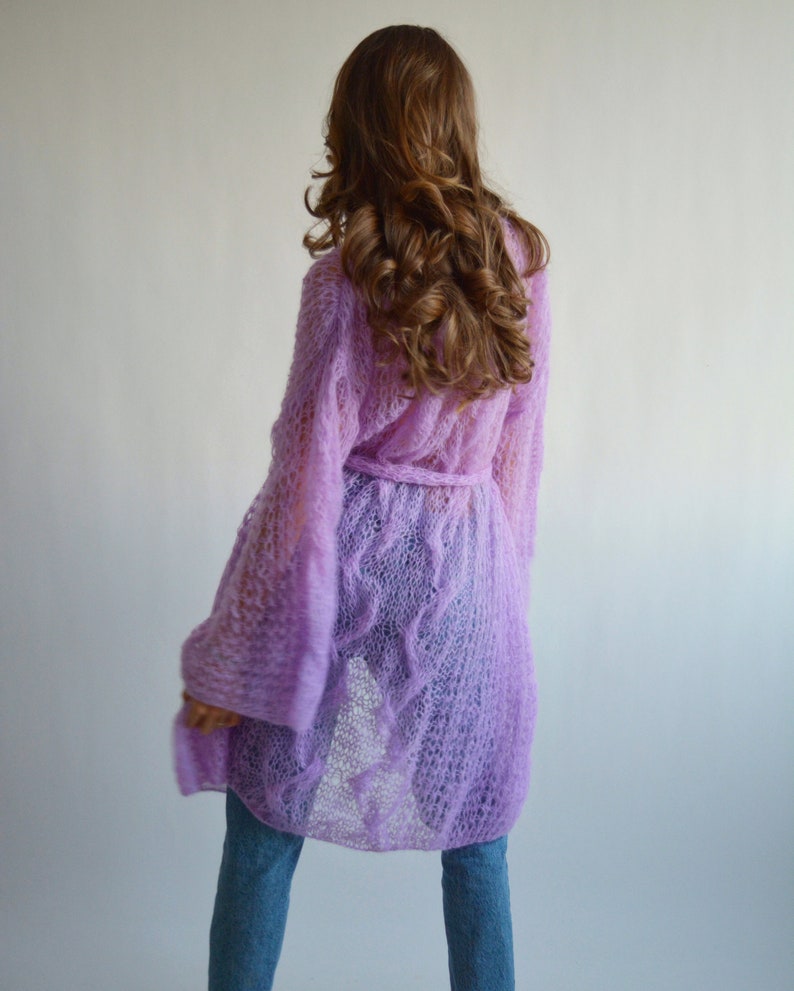 May include: A long, light purple knitted cardigan with a tie belt. The cardigan is made of a delicate, open-weave fabric.