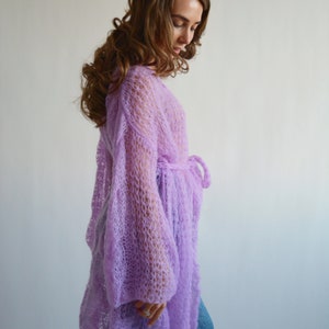 May include: A long, light purple knitted cardigan with a loose fit and long sleeves. The cardigan has a tie at the waist.