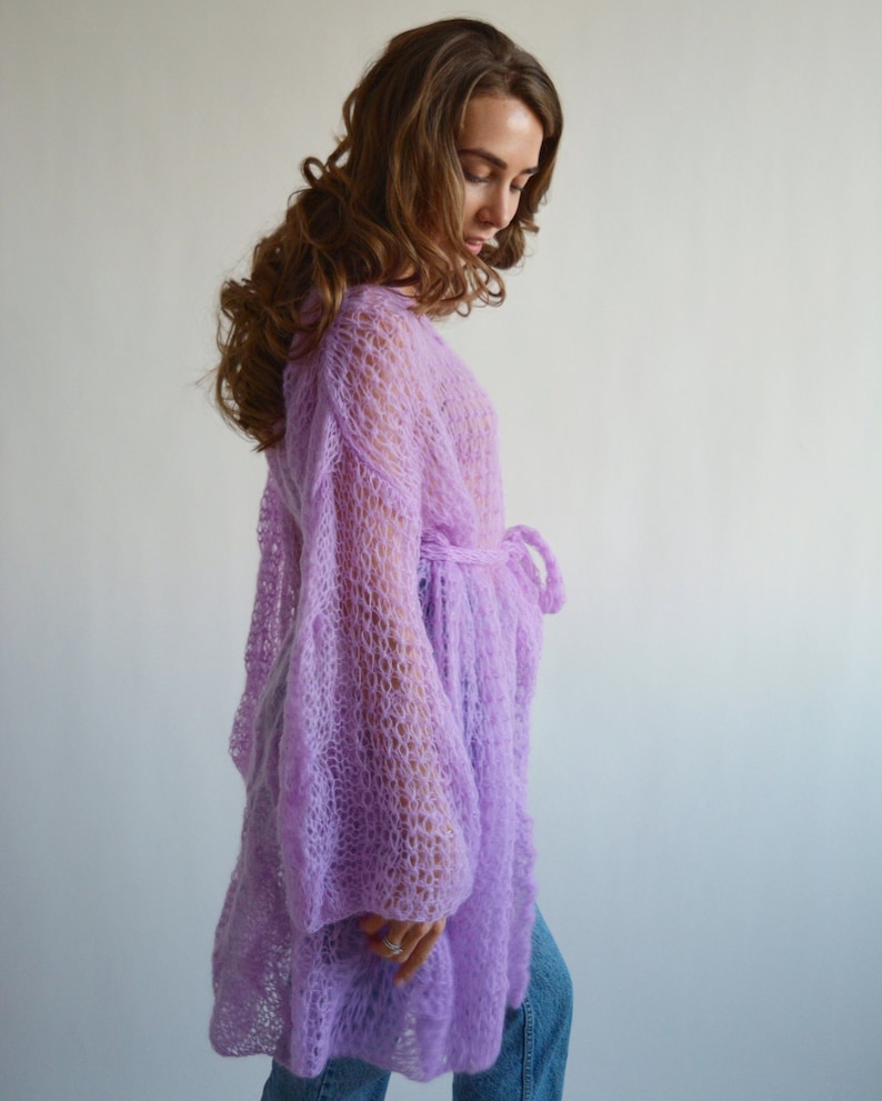 May include: A long, light purple knitted cardigan with a loose fit and long sleeves. The cardigan has a tie at the waist.