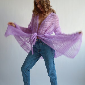 May include: A woman wearing a light purple, open-front, knit cardigan with long sleeves and a tie at the waist. She is wearing blue jeans and has her hair down.
