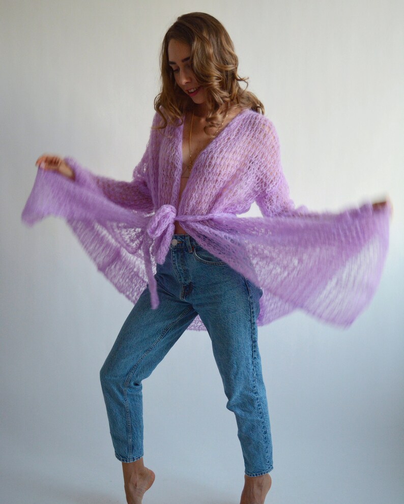 May include: A woman wearing a light purple, open-front, knit cardigan with long sleeves and a tie at the waist. She is wearing blue jeans and has her hair down.