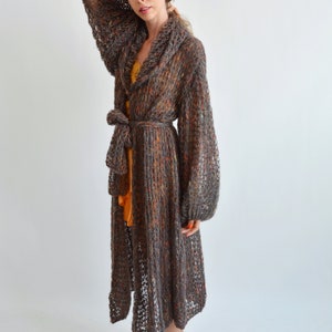 May include: A long, brown knit cardigan with a shawl collar and a tie belt. The cardigan has a loose, flowing fit and is made of a soft, lightweight yarn.