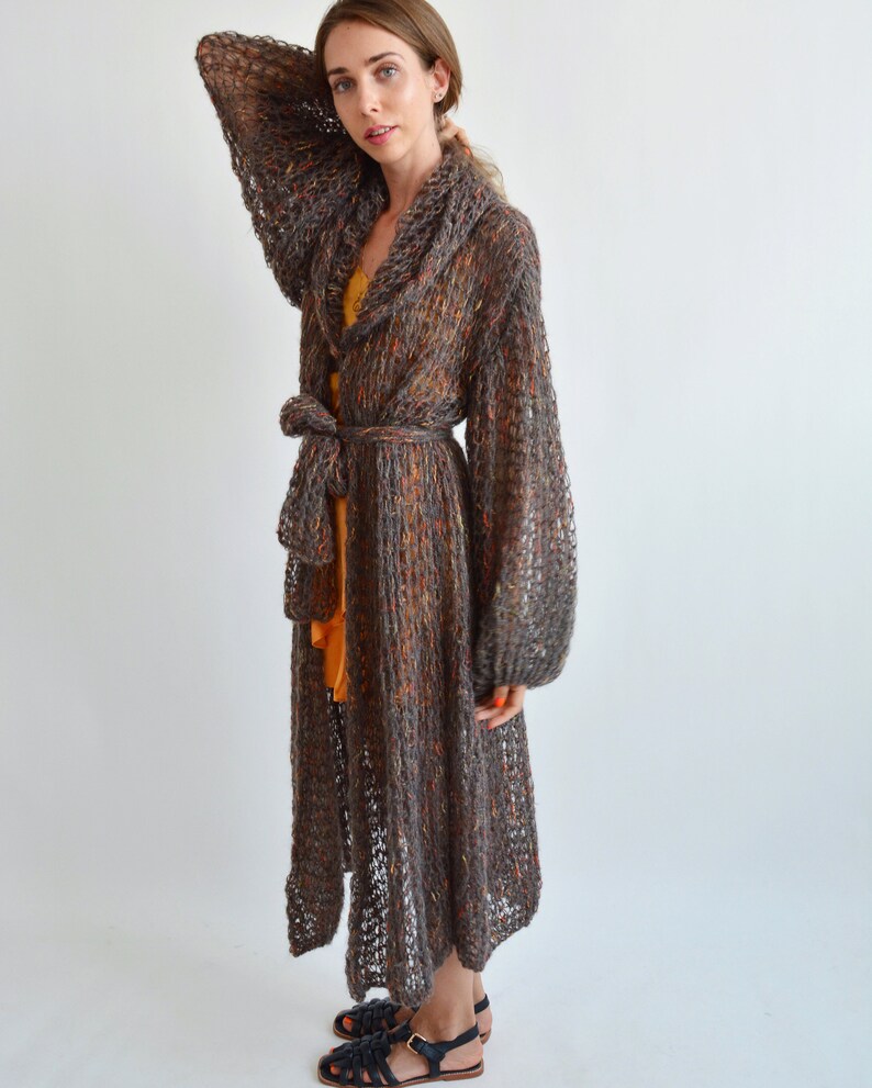 May include: A long, brown knit cardigan with a shawl collar and a tie belt. The cardigan has a loose, flowing fit and is made of a soft, lightweight yarn.