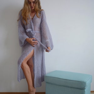 May include: A long, light purple knitted cardigan with a tie belt and a slit up the front. The cardigan is made of a soft, fuzzy yarn and has a relaxed fit.