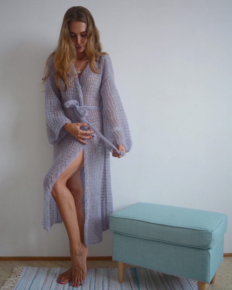 May include: A long, light purple knitted cardigan with a tie belt and a slit up the front. The cardigan is made of a soft, fuzzy yarn and has a relaxed fit.