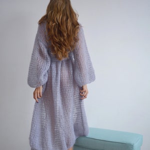 May include: A long, light purple knitted cardigan with a loose fit and long sleeves. The cardigan is made of a soft, airy yarn and has a delicate, open-weave pattern.