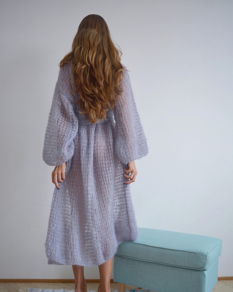 May include: A long, light purple knitted cardigan with a loose fit and long sleeves. The cardigan is made of a soft, airy yarn and has a delicate, open-weave pattern.