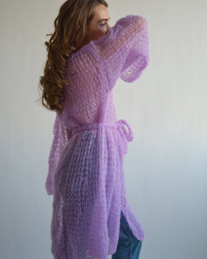 May include: A long, light purple knitted cardigan with a tie belt. The cardigan is made of a loose, open weave and has a relaxed fit.